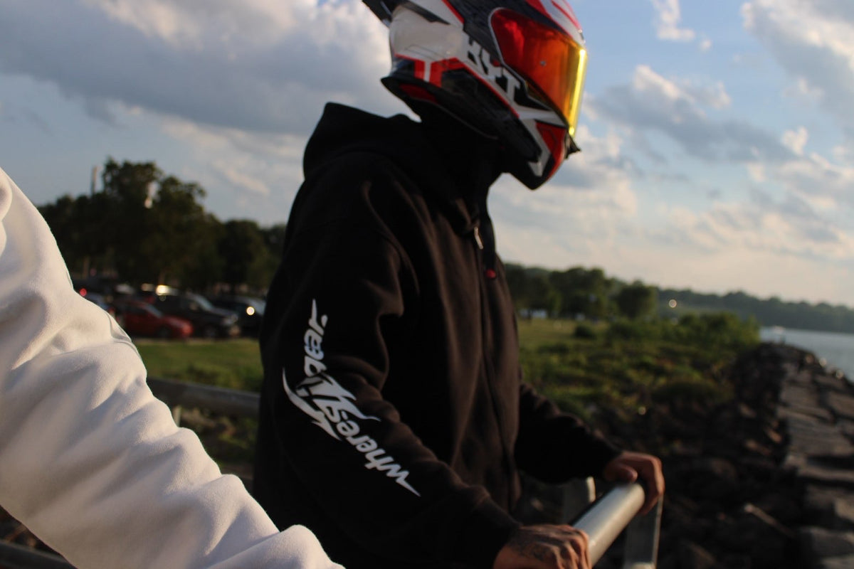 Rider in a black WHERES7THGEAR hoodie with orange visor standing by the water at sunset