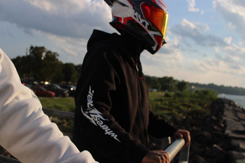 Rider in a black WHERES7THGEAR hoodie with orange visor standing by the water at sunset