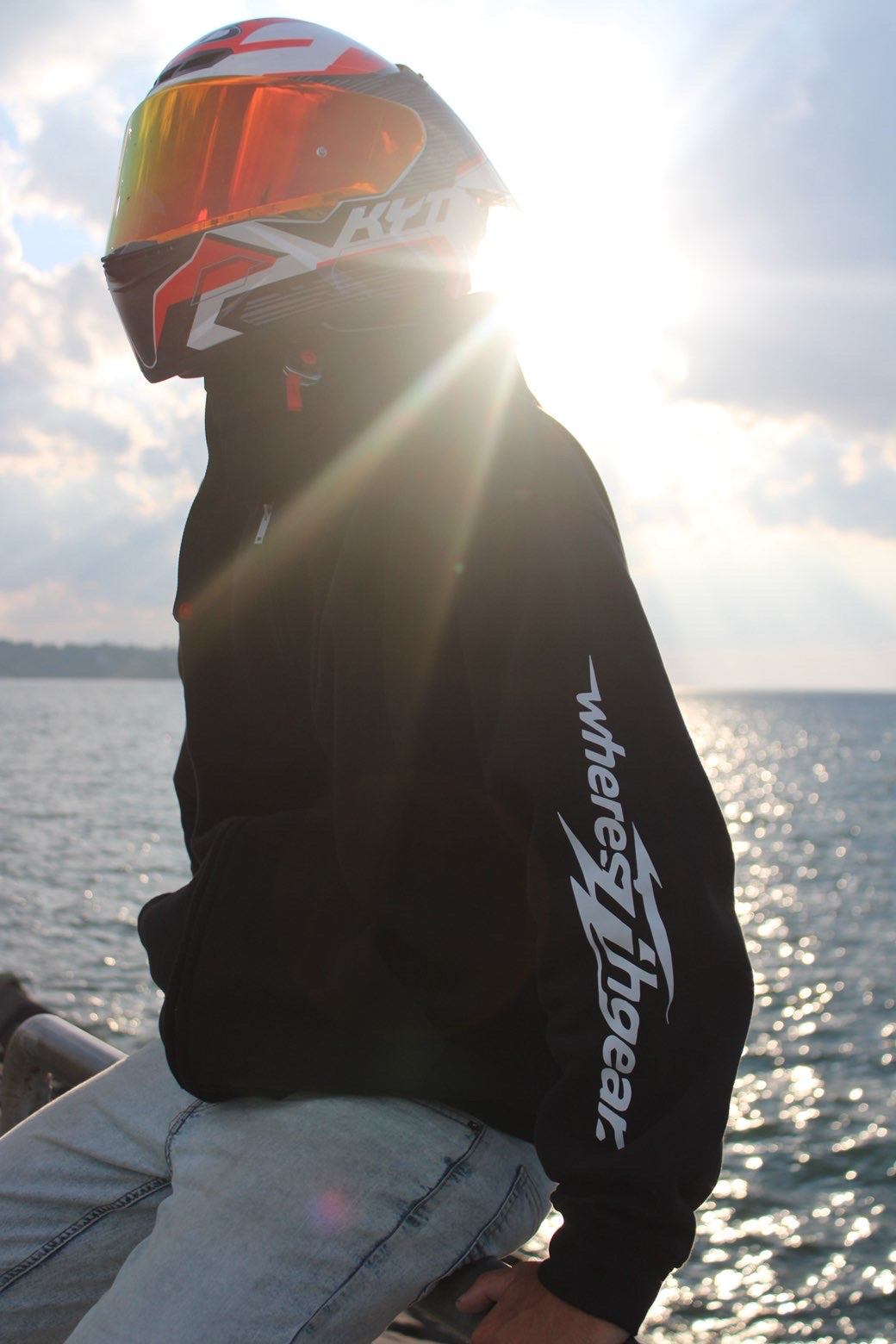 Rider in a black WHERES7THGEAR hoodie sitting by the water with sun flaring behind